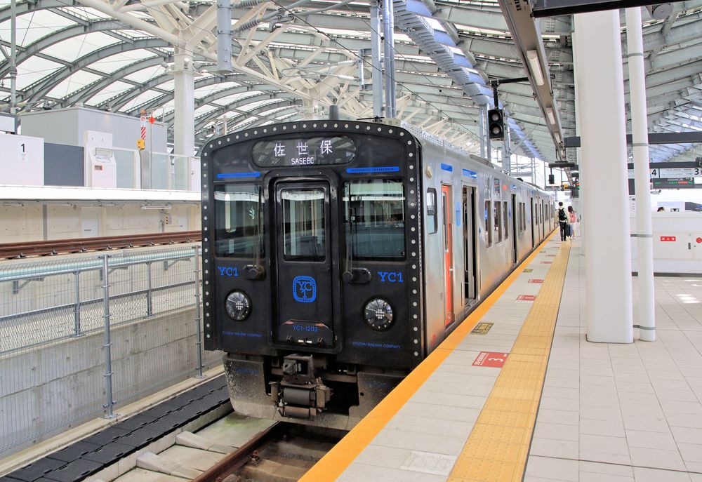 0374 Through trains to Sasebo via the Ômura-sen are now (since June 2021) worked by the new diesel-electric hybrid sets of class YC1. YC stands for "yasashikute chikaramochi" ("gentle and strong"). In the picture train set YC 1-203 is waiting at the new Nagasaki terminus for departure to Sasebo, via Utsutsugawa and the Ômura-sen. (2023)